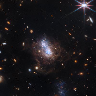 Dwarf galaxy I Zwicky 18 looks like a cluster of blue stars surrounded by brown clouds. A smaller companion galaxy is underneath